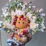 Handheld view of a handmade coastal floral pod doll with a large white-and-blue flower crown and shell-decorated wave base