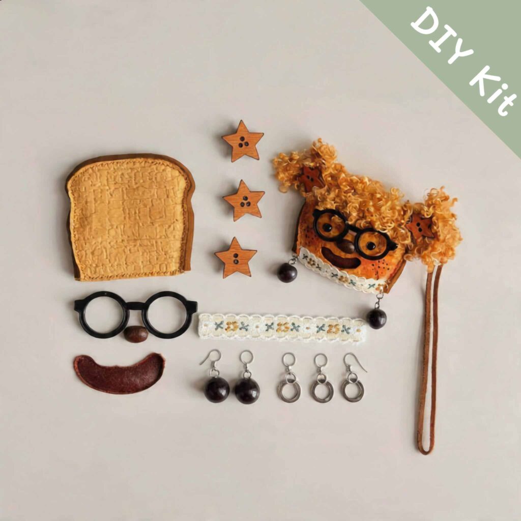 Flat lay of a DIY pod doll kit featuring a toast-shaped doll base, curly-haired face piece, glasses, star accents, lace trim, and hanging hardware