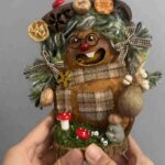 Handheld view of handmade forest granny pod doll with round glasses, mossy yarn hair, mushroom details, and a woodland display base