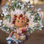 Close-up of the pod doll’s floral crown and bouquet details with layered blooms and coastal styling