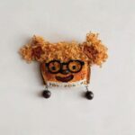 Front view of a small handmade pod doll with warm toast colors, lace trim, and dangling bead details