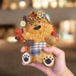 Handheld front view of a double-sided handmade pod doll with curly beard, glasses, plaid patch pocket, paw feet, and woodland-style details.