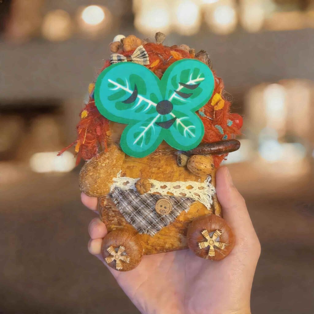 Handheld close-up of the pod doll with a magnetic green clover-style face attached, featuring curly red hair, plaid fabric details, and wooden wheel feet.