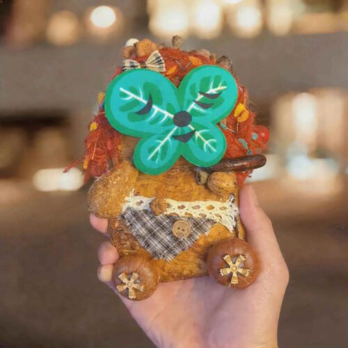Handheld close-up of the pod doll with a magnetic green clover-style face attached, featuring curly red hair, plaid fabric details, and wooden wheel feet.
