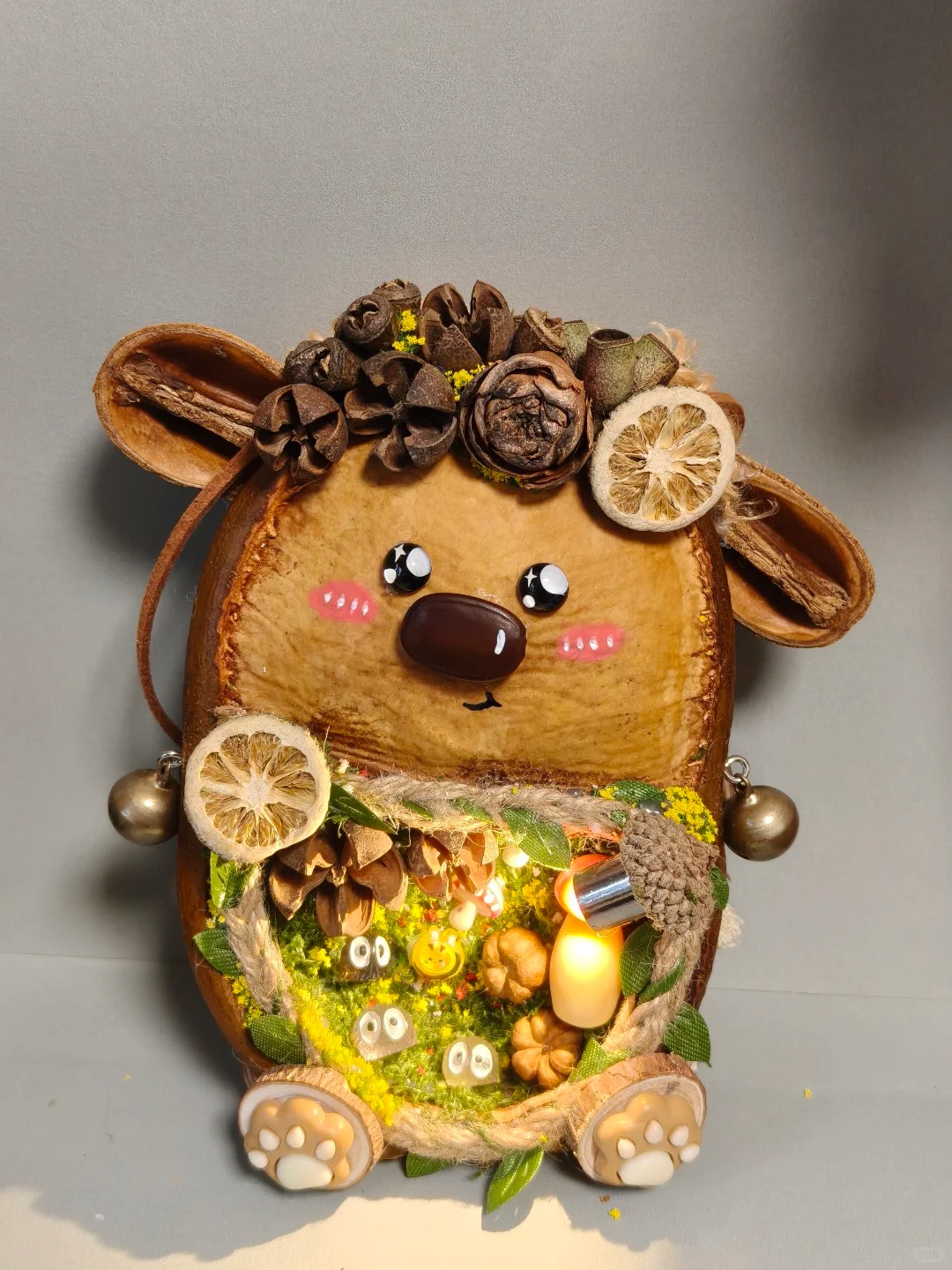 Front view of an animal-inspired Poddoll with large ears, dried citrus, flowers, paw details, bells, and an illuminated miniature scene