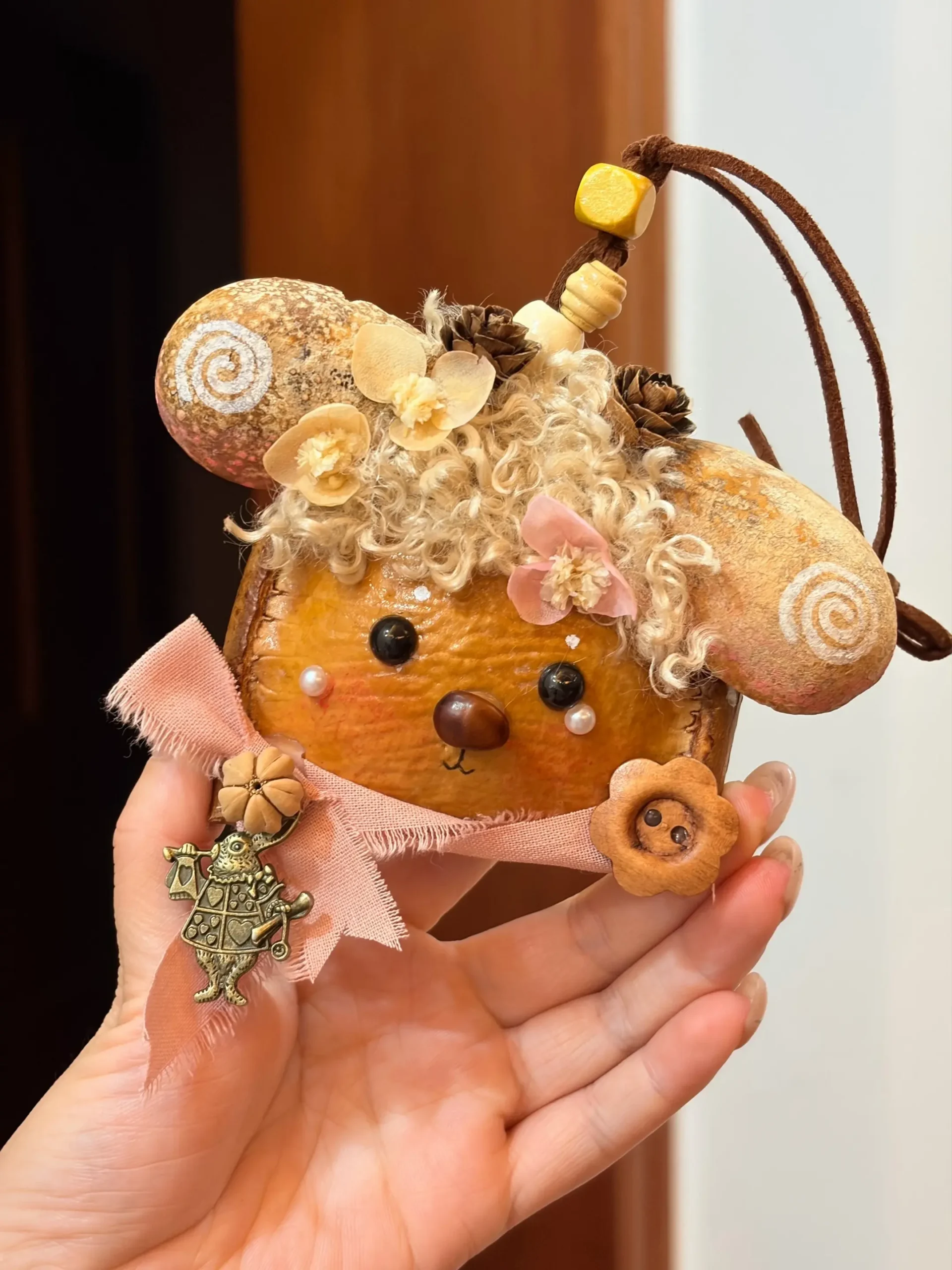 Hand holding a sheep-inspired hanging Poddoll with wide ears, pale curly hair, dried flowers, a pink scarf, and natural seed decorations