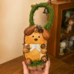 Back of a double-sided Poddoll showing a handmade dog-like character with natural ears, a blue bow, buttons, and a moss-covered handle