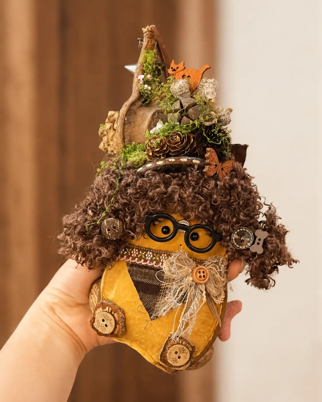 Hand holding a person-like Poddoll with round glasses, curly brown hair, fabric clothing, and a miniature woodland scene above the head