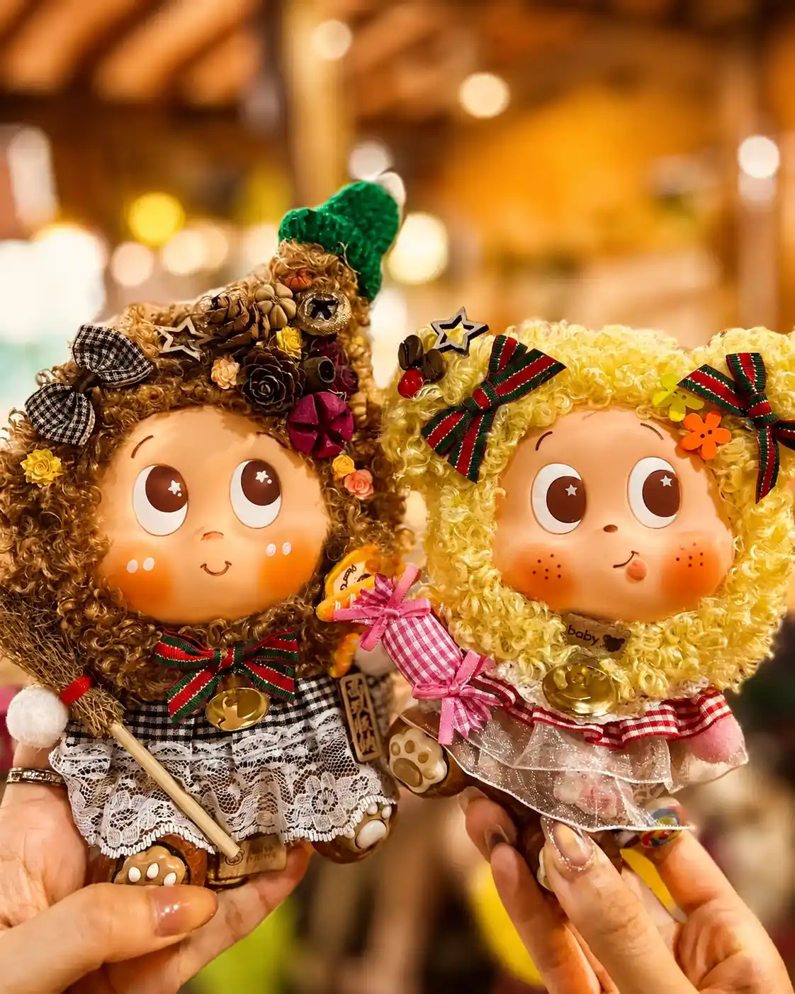 Hands holding two finished Poddolls with contrasting curly hair, lace clothing, bows, bells, and small handmade props