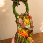 Hand holding a light-up miniature forest Poddoll with a fairy, fox, mushrooms, moss, bell, and wooden fence