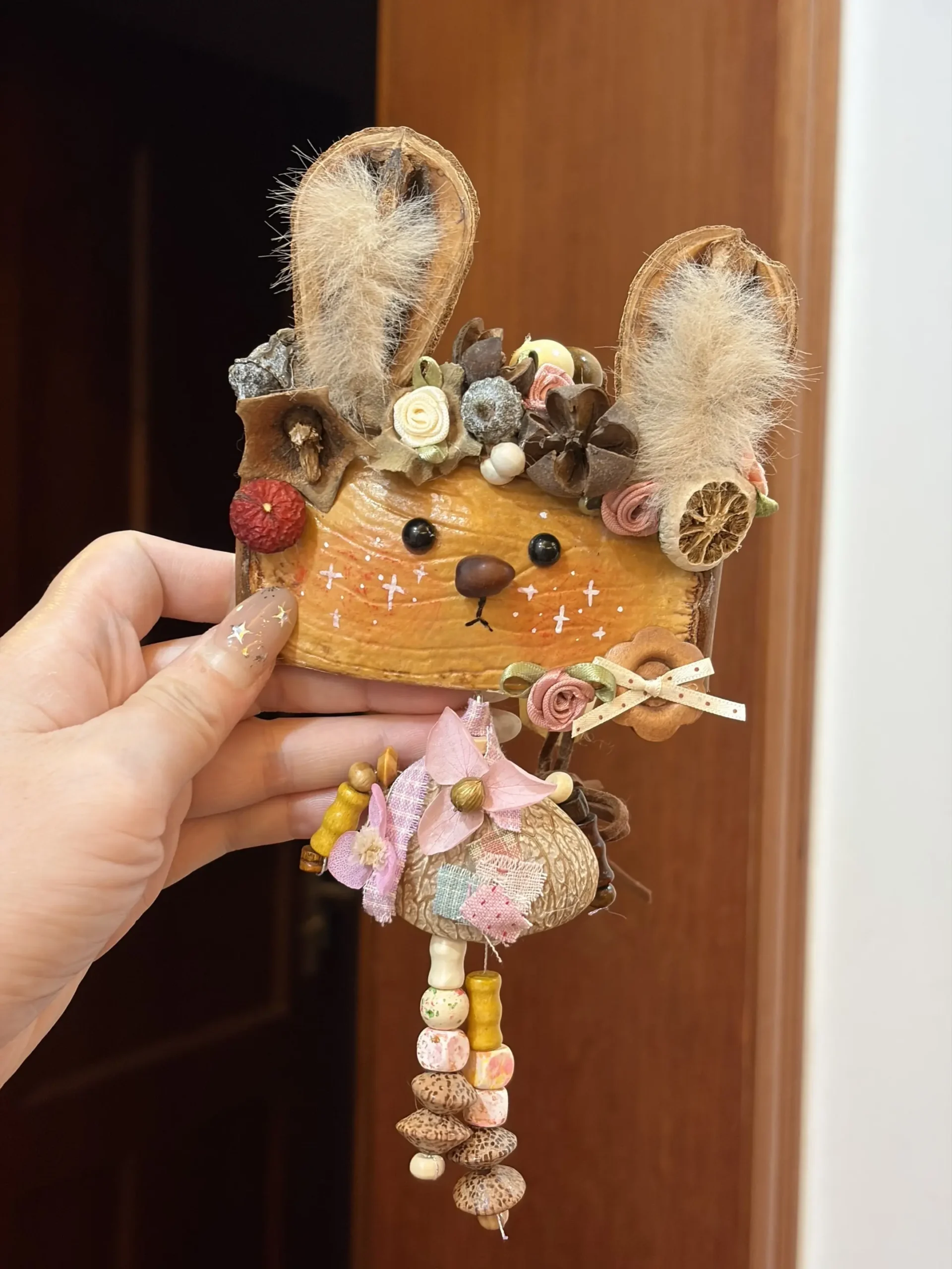 Hand holding a rabbit-inspired hanging Poddoll with tall fuzzy ears, floral details, a pink bow, and dangling natural decorations