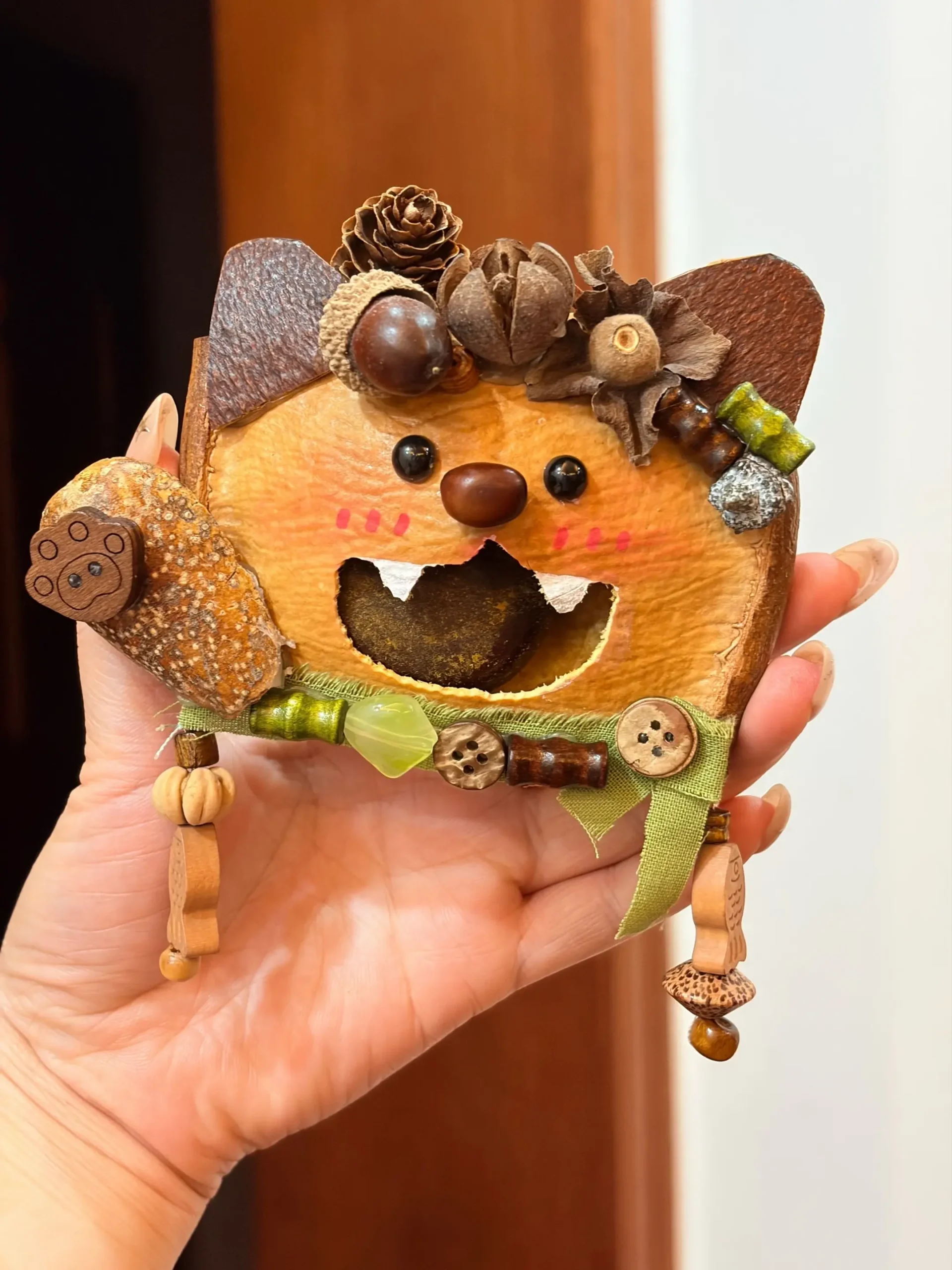 Hand holding a cat-inspired Poddoll with pointed ears, a large open mouth, natural seed decorations, buttons, and a green fabric bow