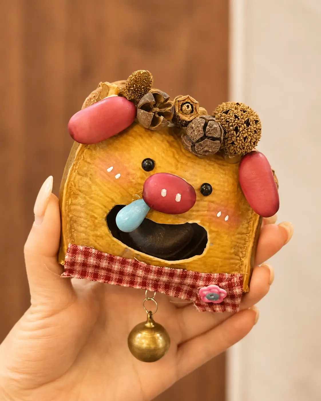 Hand holding a pig-inspired Poddoll with pink ears and snout, a large open mouth, blue tongue, natural pod decorations, plaid trim, and a hanging bell