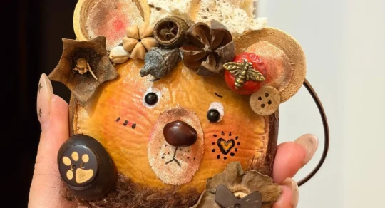 Hand holding a bear-inspired Poddoll with round ears, a tall red party hat, pinecone decorations, buttons, and natural pod details