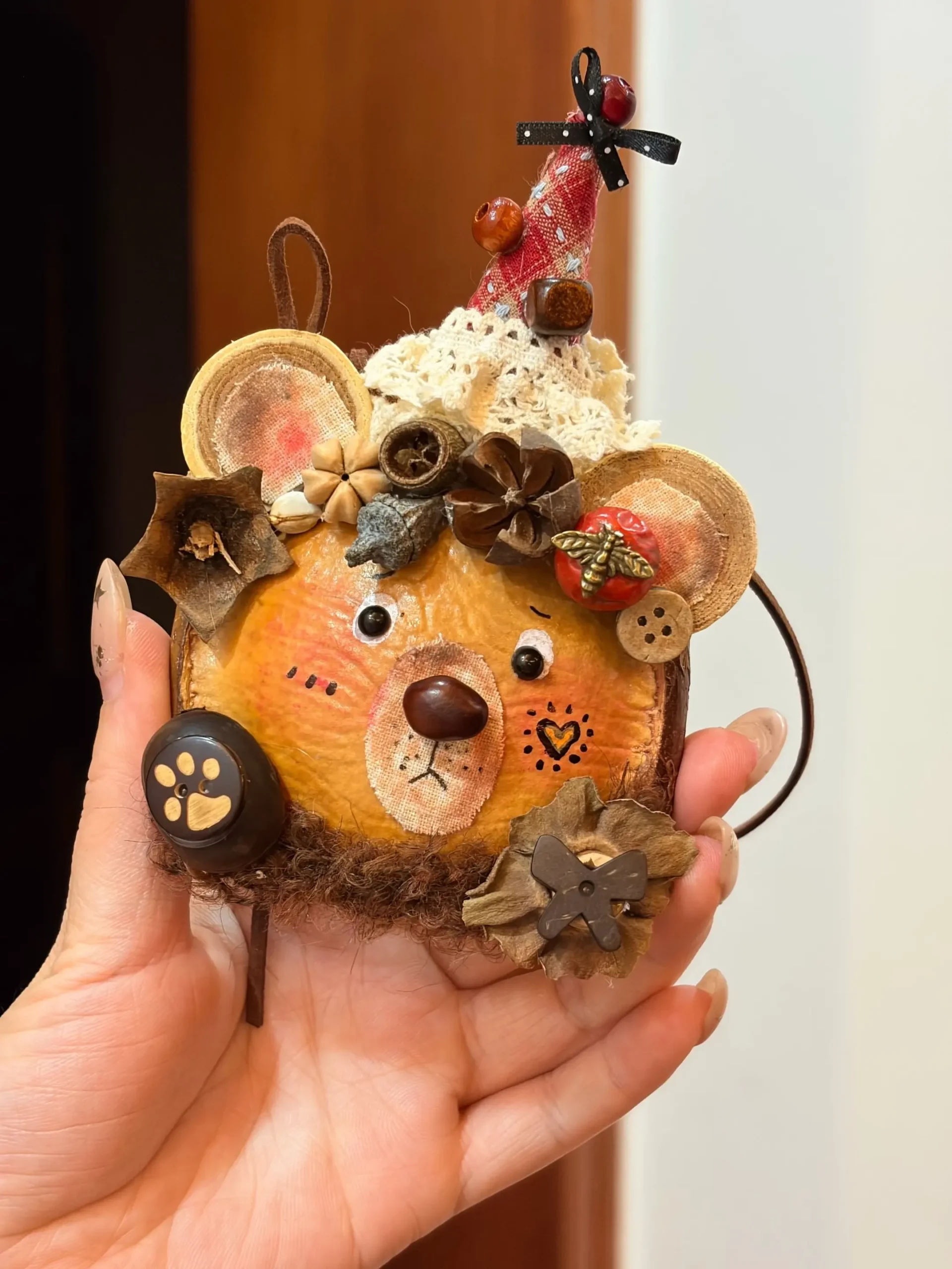 Hand holding a bear-inspired Poddoll with round ears, a tall red party hat, pinecone decorations, buttons, and natural pod details