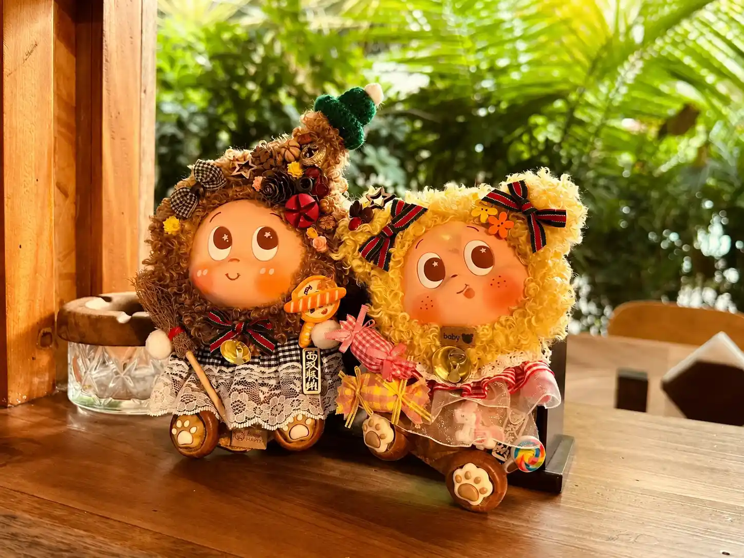 Two handmade Poddolls displayed on a wooden window shelf with curly hair, layered clothing, bows, and green plants behind them
