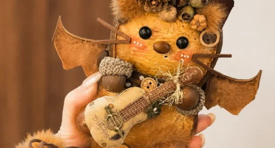 Hand holding a bat-inspired Poddoll with wide natural wings, a pointed hat, pinecone decorations, a small stringed instrument, and a bell