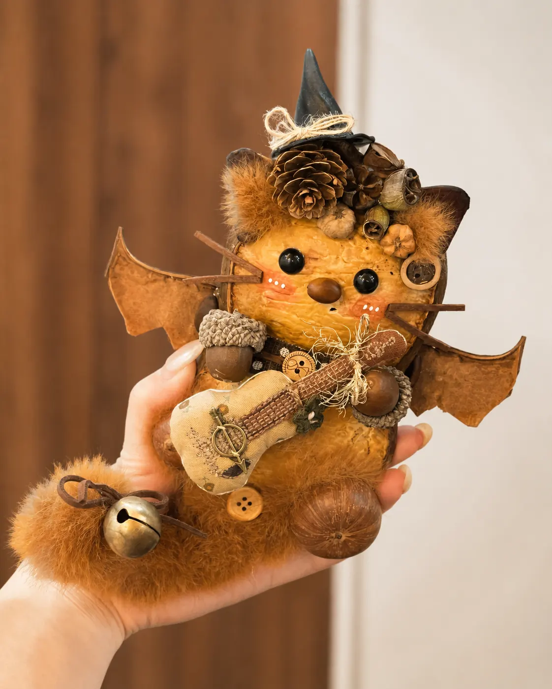 Hand holding a bat-inspired Poddoll with wide natural wings, a pointed hat, pinecone decorations, a small stringed instrument, and a bell